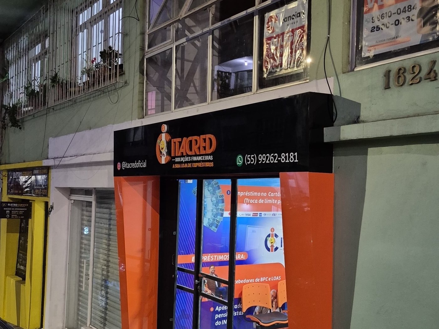 Fachada da Itacred à noite com a testeira iluminada e a vitrine acesa, ao lado de lojas já fechadas